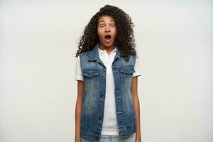 Shocked young dark skinned brunette curly female looking amazedly to camera with wide eyes and mouth opened, keeping hands along body while posing over white background photo