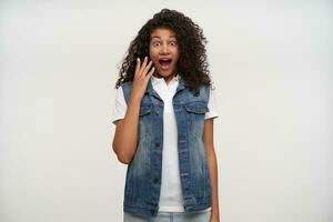 Studio photo of open-eyed pretty dark skinned curly woman raising palm to her face and looking surprisedly at camera, dressed in jeans vest and white shirt over white background