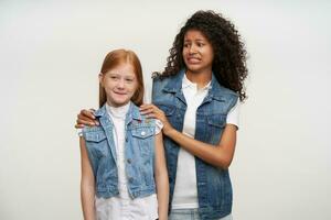 Discouraged young dark skinned brunette lady with long curly hair twisting mouth and frowning eyebrows while looking at lovely female kid with foxy hair in front of her, isolated over white background photo