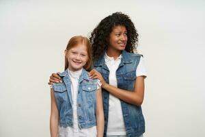 Lovely redhead long haired little girl in casual clothes looking cheerfully to camera and smiling widely while standing in front of confused dark skinned curly woman, isolated over white background photo