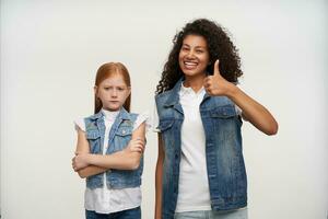 Indoor shot of cheerful pretty young curly brunette lady with dark skin showing raised thumb and smiling joyfully to camera while posing over white background with foxy long haired upset female kid photo