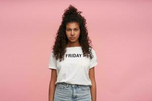 Attractive young curly long haired brunette female with dark skin keeping hands along her body while posing over pink background, looking at camera with calm face and folded lips photo