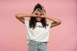 Portrait of cheerful funny curly brunette lady with dark skin making glasses from raised hands while standing over pink background, being in high spirit and smiling widely photo