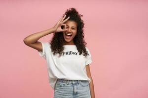 Joyful pretty young brunette dark skinned woman with curly long hair standing over pink background with raised hand, folding ok gesture and smiling widely to camera, dressed in casual clothes photo