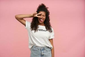 Shot of funny young curly long haired brunette woman with dark skin closing one eye and showing tongue to camera, folding victory sigh and keeping it near her face, isolated over pink background photo