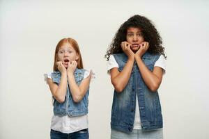 Frightened open-eyed young girls wearing family look while standing over white background, holding faces with hands and looking afraid at camera with raised eyebrows photo