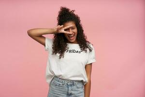 Indoor shot of happy young brunette dark skinned female with curly long hair dressed in white t-shirt and blue jeans posing over pink background, raising peace gesture to her face and smiling joyfully photo