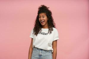 Joyful beautiful young dark skinned brunette woman with long curly hair wearing casual hairstyle while posing over pink background, looking cheerfully aside and laughing with wide mouth opened photo