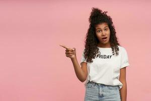 Indoor shot of bewildered young curly long haired brunette lady with dark skin pointing aside with index finger and looking confusedly to camera with raised eyebrows, isolated over pink background photo