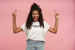 Joyful attractive young curly brunette lady with dark skin crossing fingers for good luck and looking cheerfully to camera with broad sincere smile,wearing white t-shirt and jeans over pink background photo