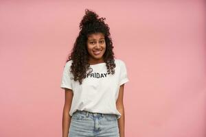 Confused dark skinned curly brunette female looking at camera with oops face and biting underlip, keeping hands along body while posing over pink background, dressed in casual clothes photo