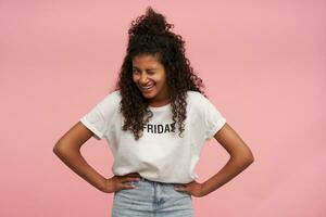 Joyful pretty young curly brunette lady with dark skin holding hands on her waist and laughing happily, giving wink to camera while standing over pink background photo