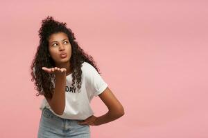 Indoor shot of attractive young curly dark skinned brunette female folding lips in air kiss and keeping palm raised, wearing white t-shirt and blue jeans while posing over pink background photo