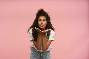 Portrait of pleasant looking young curly brunette dark skinned lady with casual hairstyle raising palm up and blowing air kiss to camera, posing over pink background inwhite t-shirt and blue jeans photo