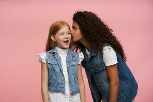 Horizontal shot of pretty young dark skinned brunette woman with long curly hair telling secret story to excited little red haired girl, wearing family look while posing over pink background photo