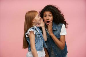 Indoor photo of attractive young girls wearing family look while posing over pink background, keeping hands near their faces while sharing with each other surprising news