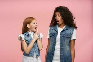 Indoor photo with young couple of shocked pretty girls looking amazedly on each other with wide mouth opened and rounding surprisedly eyes, isolated over pink background