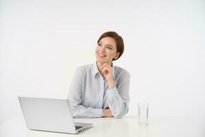 Happy young lovely brunette woman with casual hairstyle working in modern office with laptop, smiling pleasantly while looking aside and holding chin with raised hand, isolated over white background photo