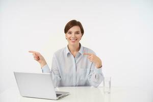 Cheerful young pretty short haired brunette woman working in office with her laptop and looking positively at camera with wide smile, poiting aside with forefingers while sitting over white background photo