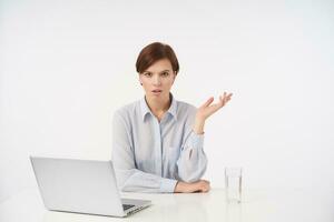 Serious young pretty brown haired female with short trendy haircut frowning scowlingly her eyebrows and keeping palm raised while sitting at table over white background photo