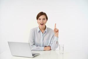 Joyful young pretty short haired brunette female with casual hairstyle sitting over white background and showing upwards with raised index finger, looking cheerfully at camera with wide mouth opened photo