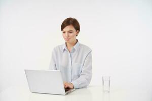 Indoor photo of charming young short haired brunette lady with casual hairstyle holding her hand on keyboard and typing text on laptop, isolated over white background