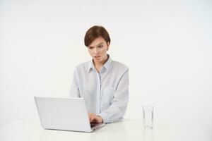 Confused young attractive short haired brunette woman with casual hairstyle keeping hand on keyboard of laptop while looking perplexedly at screen with raised eyebrow, isolated over white background photo