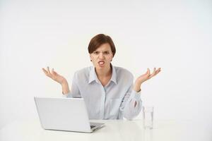 Irritated young brown haired lady with short trendy haircut frowning her face and twisting mouth while shrugging with raised palms, dressed in blue shirt while posing over white background photo