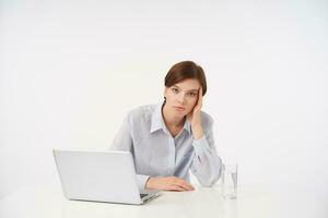 Exhausted young pretty short haired brunette lady with natural makeup leaning her head on raised hand and looking tiredly at camera, isolated over white background photo