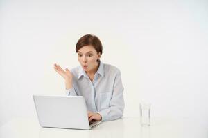 Surprised young lovely short haired brunette woman sitting at table over white background with modern laptop and looking at screen with wide eyes opened, dressed in formal clothes photo