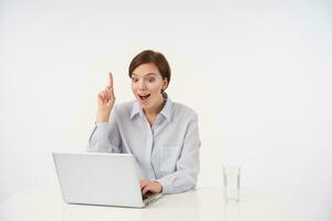 Emotional young brown haired attractive woman with casual hairstyle raising excitedly forefinger while having good idea, wearing blue shirt while posing over white background photo