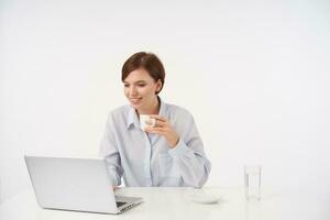 Good looking young positive short haired brunette female with natural makeup drinking cup of coffee while working at office with her laptop, isolated over white background photo