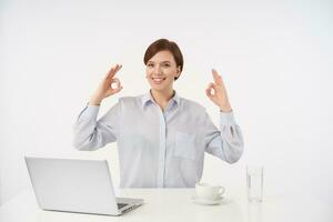 Happy young brown-eyed short haired brunette lady with casual hairstyle raising hands with ok gesture and looking positively at camera with charming smile, isolated over white background photo