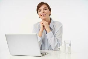 Cheerful young charming brown haired woman with short trendy haircut smiling pleasantly at camera and keeping hands under her chin, sitting over white background photo