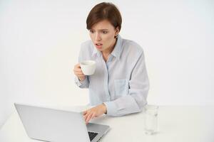 Open-eyed young pretty brown haired woman with casual hairstyle holding ceramic cup and looking amazedly at screen of laptop, reading unexpected news, isolated over white background photo
