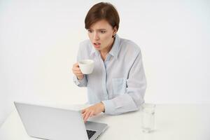 con los ojos abiertos joven bonito marrón peludo mujer con casual peinado participación cerámico taza y mirando asombrado a pantalla de computadora portátil, leyendo inesperado noticias, aislado terminado blanco antecedentes foto