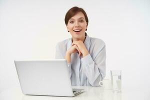 Excited young pretty short haired brunette female dressed in formal clothes folding raised hands and looking happily at camera while posing over white office background photo