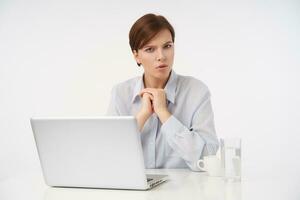 Severe young pretty short haired brunette woman with natural makeup looking seriously at camera and frowning eyebrows, folding raised hands while sitting over white background photo