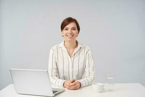 Happy young brown-eyed charming short haired brunette lady keeping her hands on countertop while sitting at table over white background, looking cheerfully at camera with broad smile photo