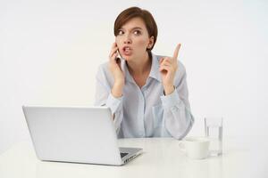 Indoor photo of young attractive brunette lady with short trendy haircut keeping her forefinger raised while having tense phone conversation, wearing blue shirt while posing over white background