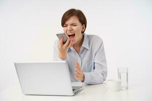 Stressed young short haired brunette woman frowning her face with closed eyes while shouting angrily into handset, having bad day at work, isolated over white background photo