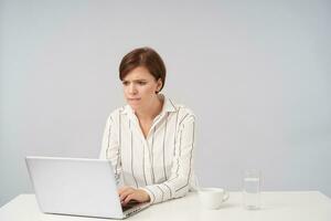 Exited young pretty brown haired lady with short trendy haircut keeping her hands on keyboard and looking worringly at screen of her laptop, sitting over white background photo