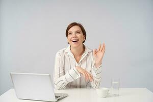 Joyful young brown haired pretty female with short trendy haircut keeping her palm raised while looking happily upwards, dressed in striped shirt while sitting over white background photo