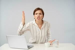 Portrait of agitated young brown haired woman with natural makeup raising emotionally hand and showing her teeth while looking fiercely at camera, sitting at table over white background photo