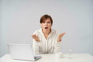 Perplexed young brown haired woman with short trendy haircut raising excitedly hands while frowning her eyebrows with opened mouth, sitting over white background photo