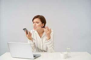 Stressed young brown haired pretty business woman having hard day at her work, holding mobile phone in raised hand and looking angrily on it, isolated over white background photo