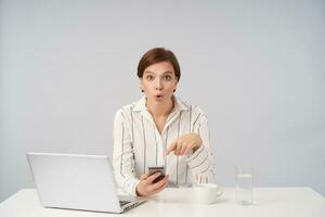 Surprised young lovely short haired brunette lady dressed in striped shirt showing on smartphone in her hand and rounding her brown eyes while looking amazedly at camera, sitting over white background photo