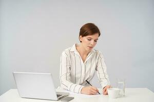 Busy young pretty short haired brunette female with casual hairstyle holding pen in her hand while making notes before important meeting, wearing white striped shirt over white background photo