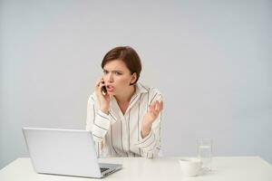 Indoor shot of young attractive short haired brunette lady with natural makeup having tense phone conversation while sitting over white background with raised palm photo