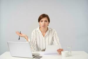 Embarrassed young short haired brunette female with natural makeup can't figure out with documents and looking confusedly at camera, frowning eyebrows with raised palm over white background photo